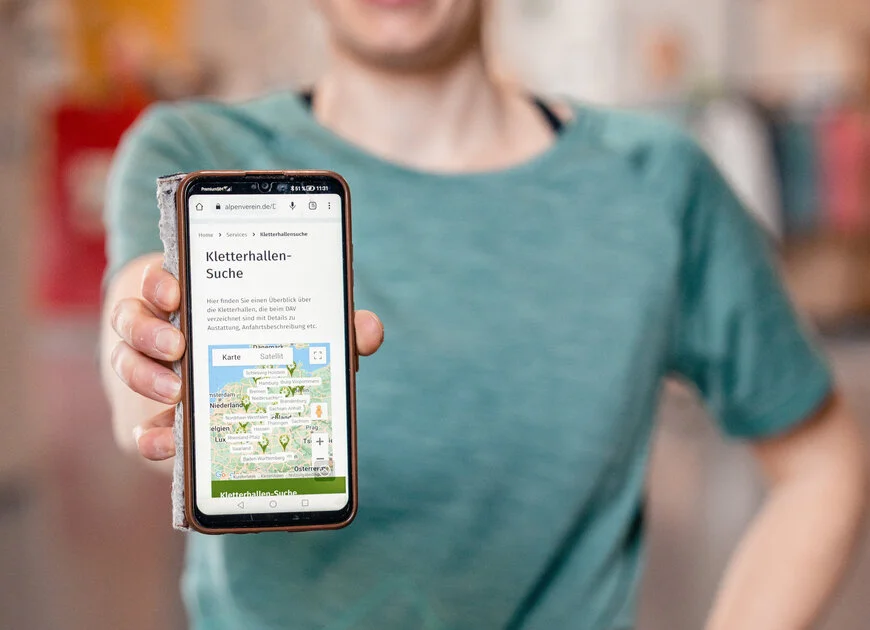 Eine Frau hält ihr Smartphone in die Kamera - auf dem Display sieht man die Kletterhallensuche | © DAV/Marisa Koch